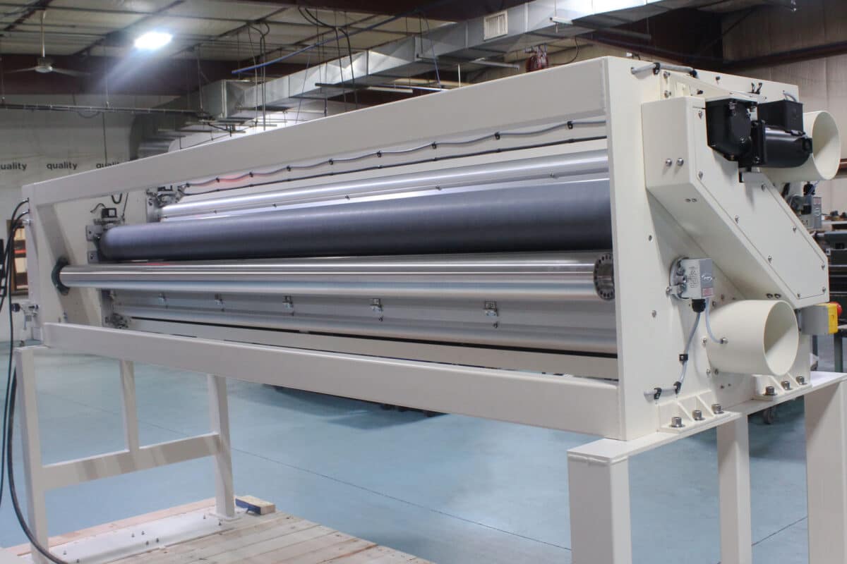 Industrial textile or paper machine with multiple rollers and a large white frame in a factory setting.