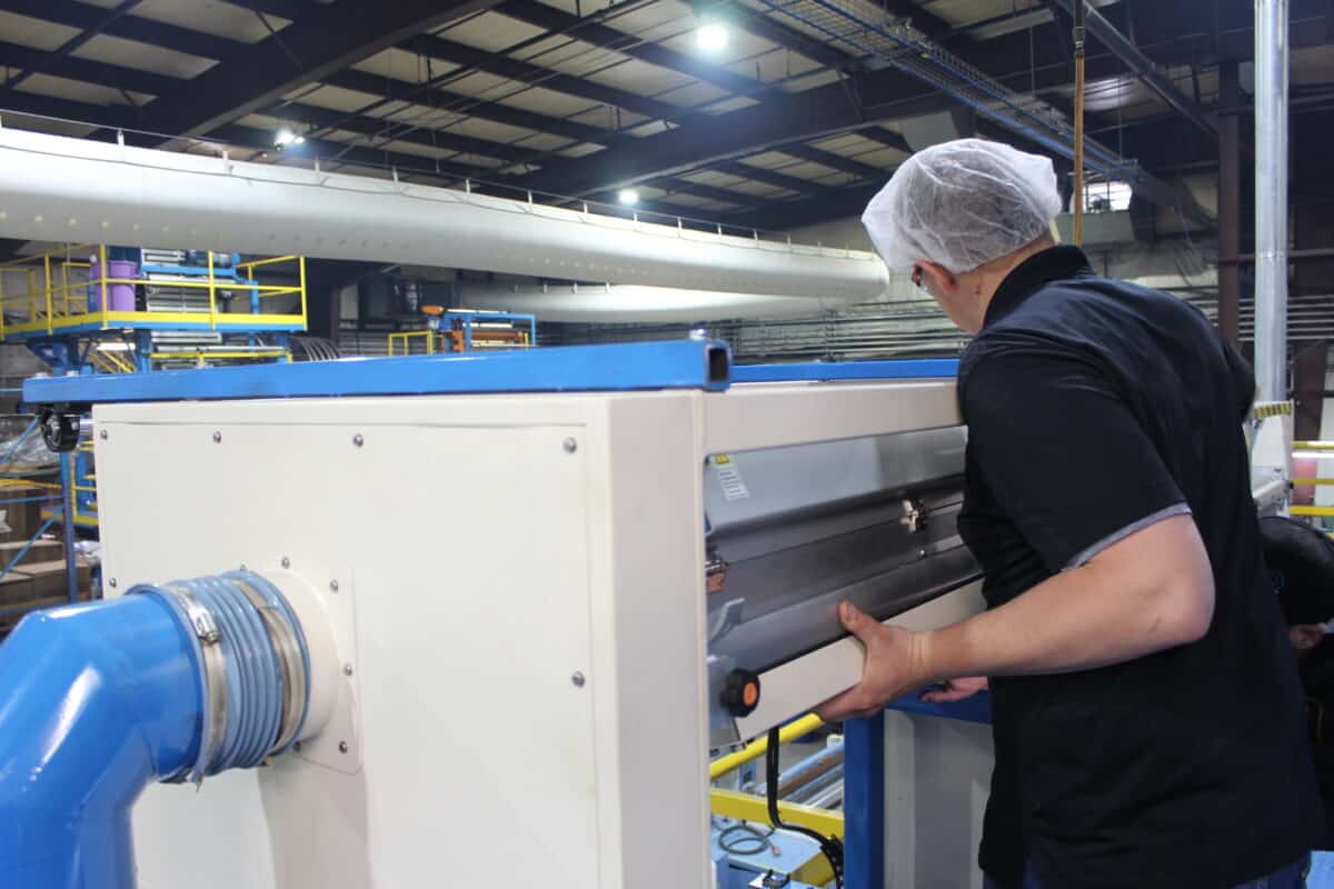 Worker in a hairnet operating a large industrial machine in a factory, with blue piping and yellow safety rails in the background.